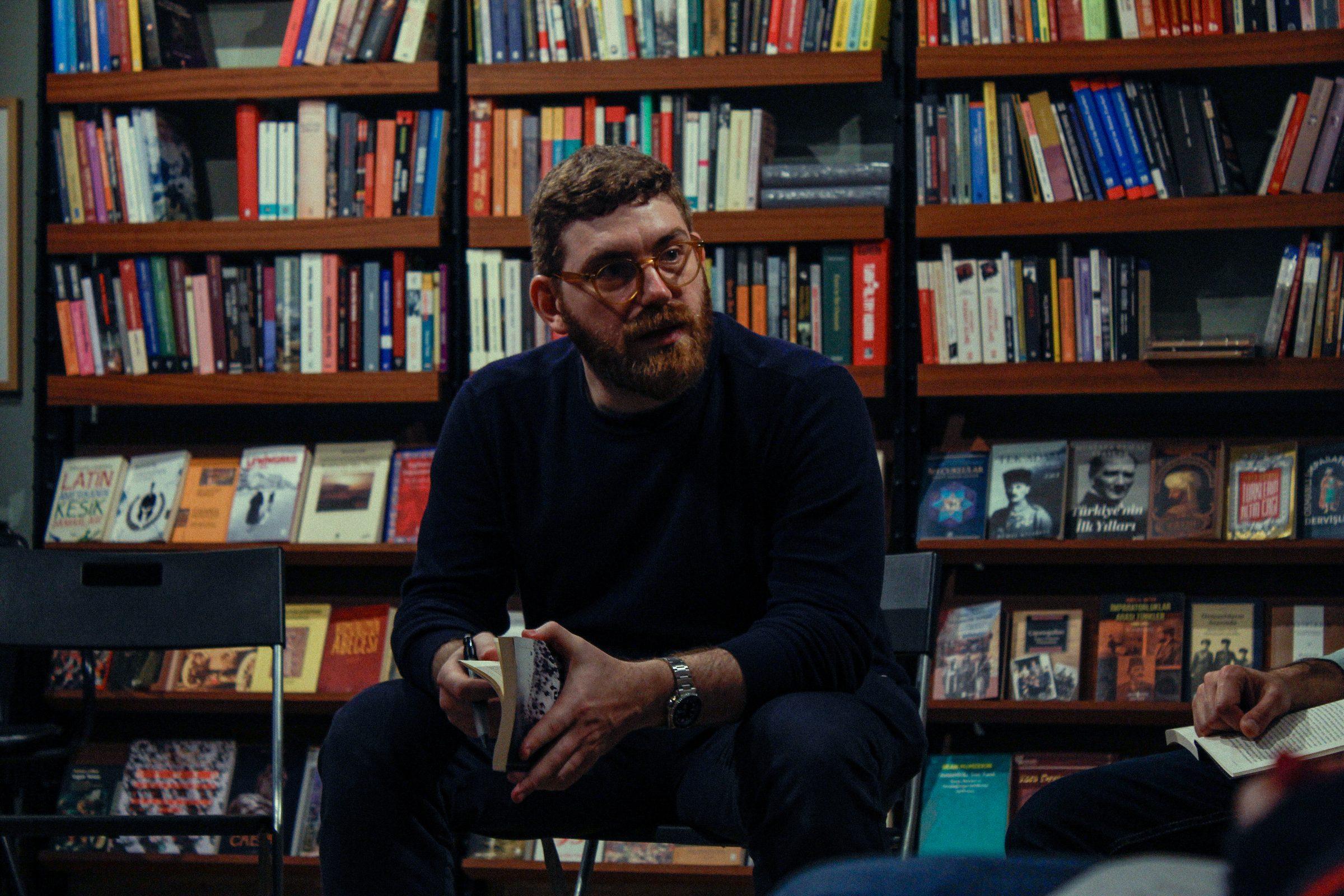 Moderatör Seyfeddin Başsaraç kitap rafları önünde konuşurken